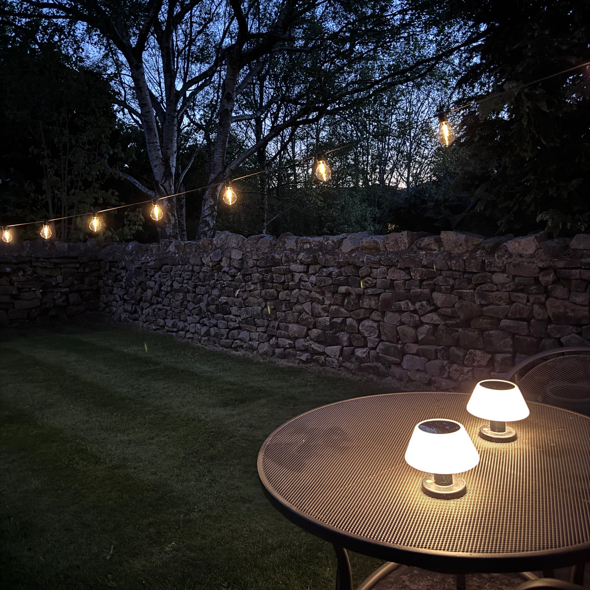 String lights on a stone wall with a tree and outdoor furniture in the background