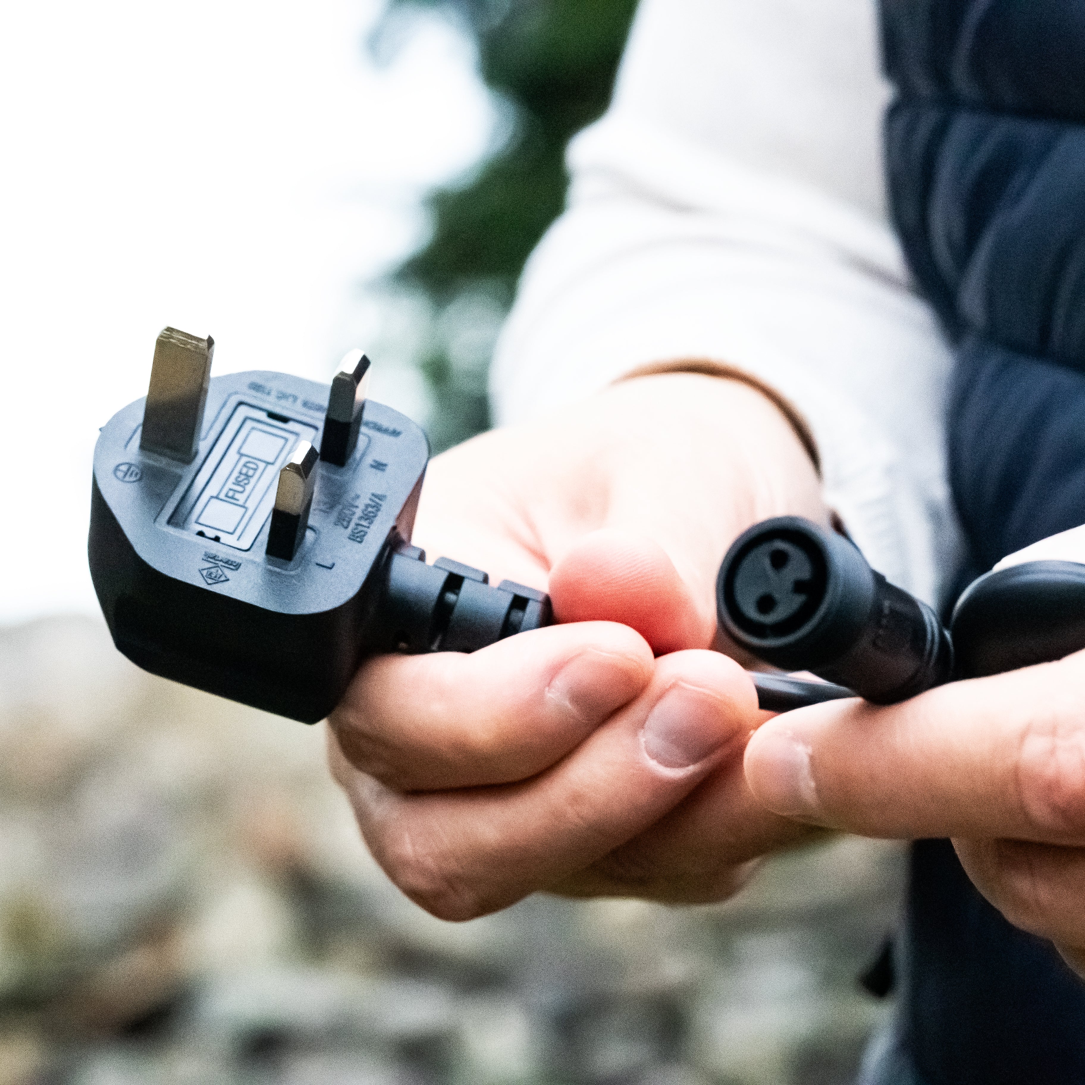 Person holding a British plug adapter with a European plug, outdoors.