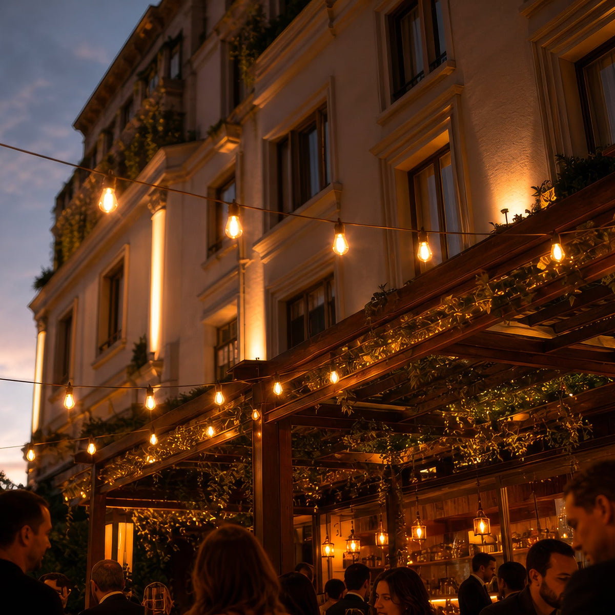 Outdoor evening scene at a building entrance with warm string lights hanging above a wooden pergola, people gathered in the foreground, and the lit facade of a classical-style building in the background.