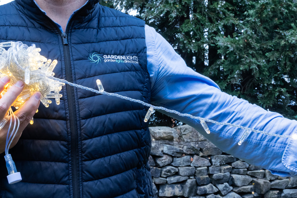 A person outdoors wearing a navy puffer vest with the Garden Light brand logo, holding a bundle of warm white fairy lights in one hand and stretching a string of clear LED lights across the frame with the other arm. A stone wall and green trees are in the background.