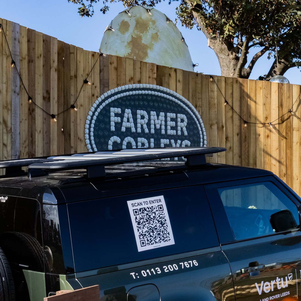 A black Vertu Land Rover SUV parked outdoors in front of a wooden fence with a large 'Farmer Copleys' sign and a moon decoration with portofino festoon lights above it. The vehicle has 'Vertu Farmer Copleys' branding on the side.