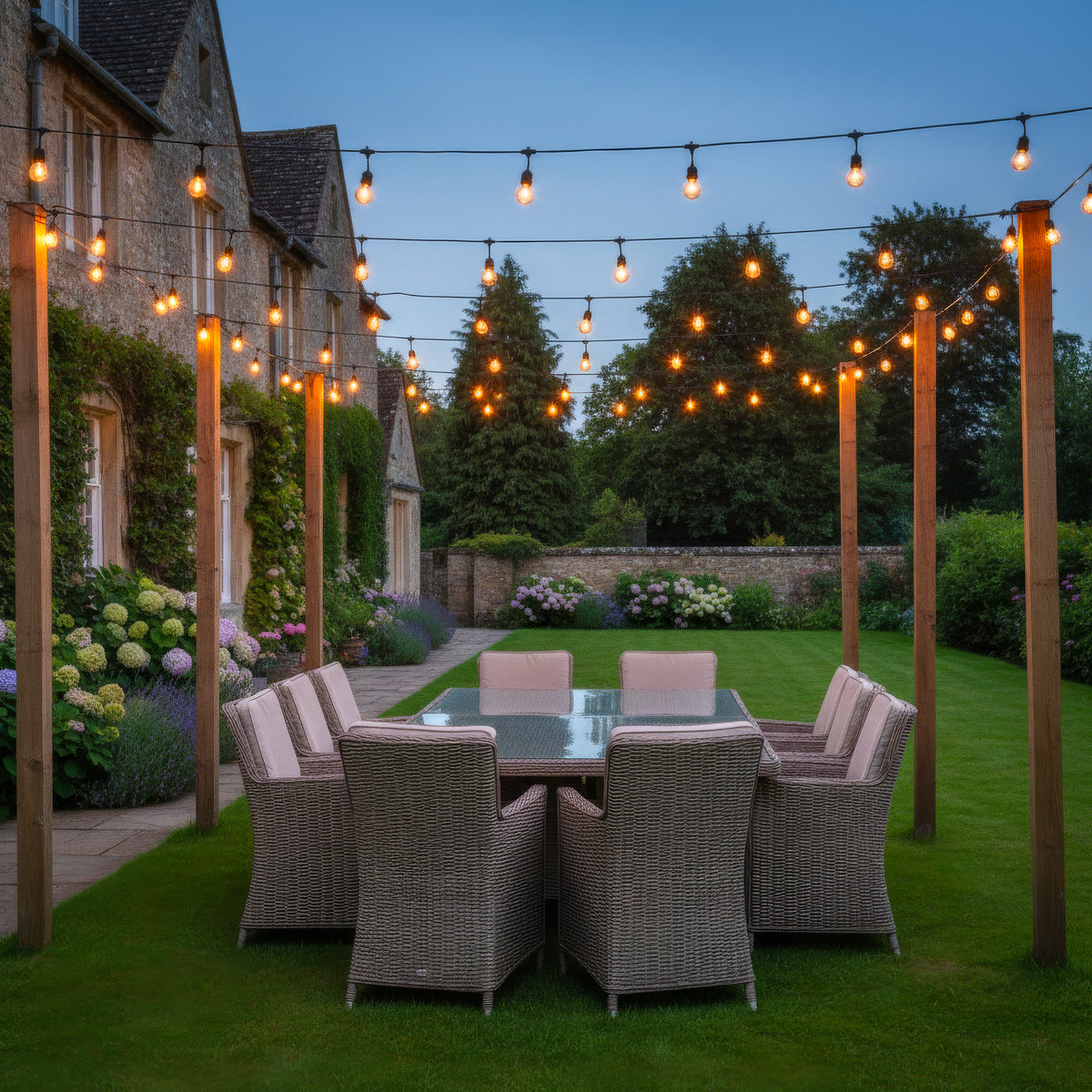 Outdoor patio dining setup with a rectangular glass-top table and six woven wicker chairs with light beige cushions, placed on a green lawn beside a stone house. Warm string lights hang overhead, with trees, flowers, and a stone wall in the background at dusk.